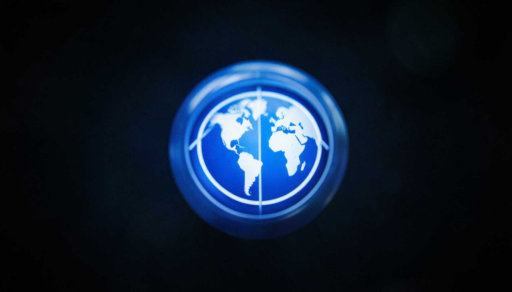 A wide cinematic frame with shallow depth of field showing an abstract open-access publishing badge with a globe motif in deep blue and white on a dark background